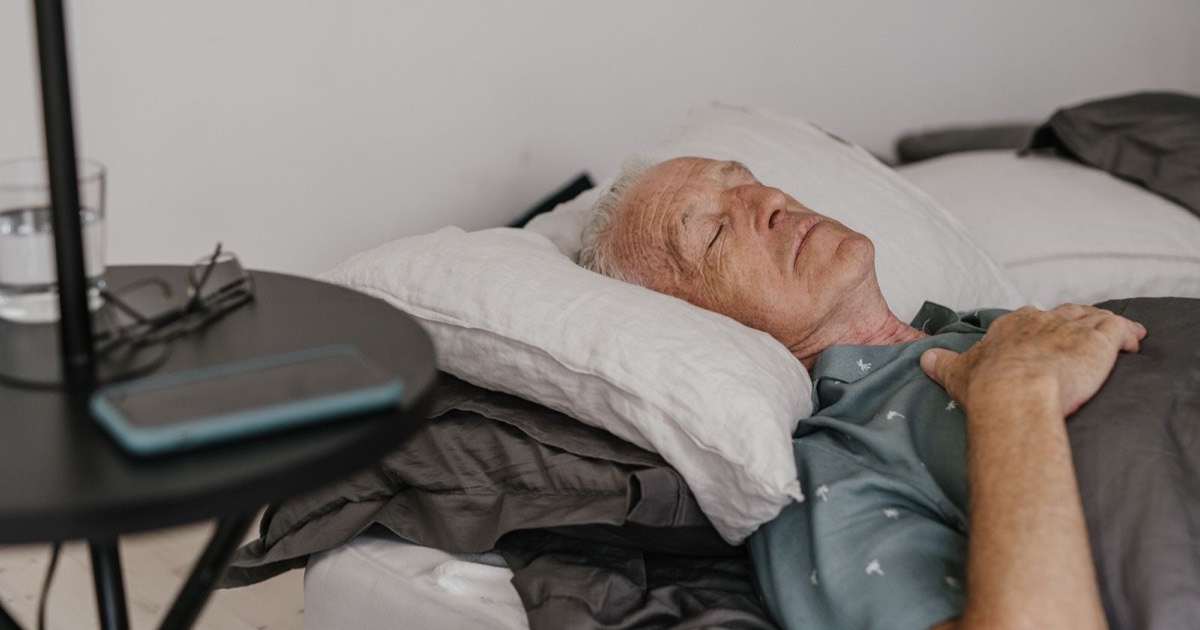 Older man asleep in bed at home beside a nightstand with a glass of water and reading glasses