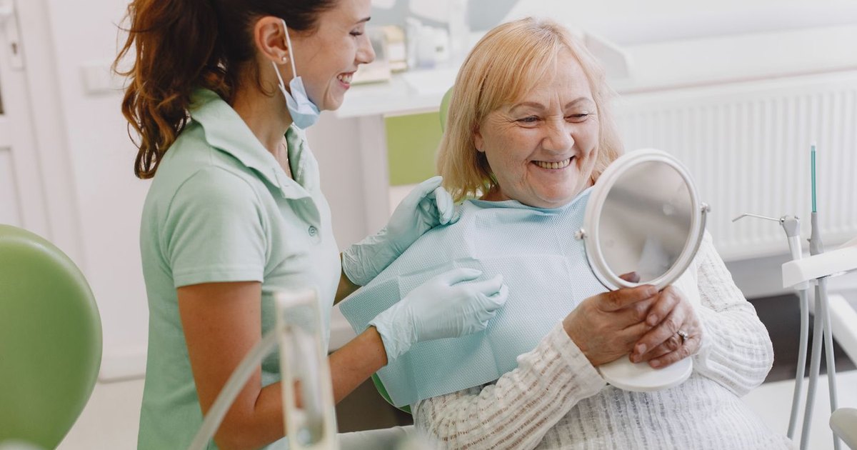 Older woman smiling at her reflection in a hand mirror while a dental hygienist looks on