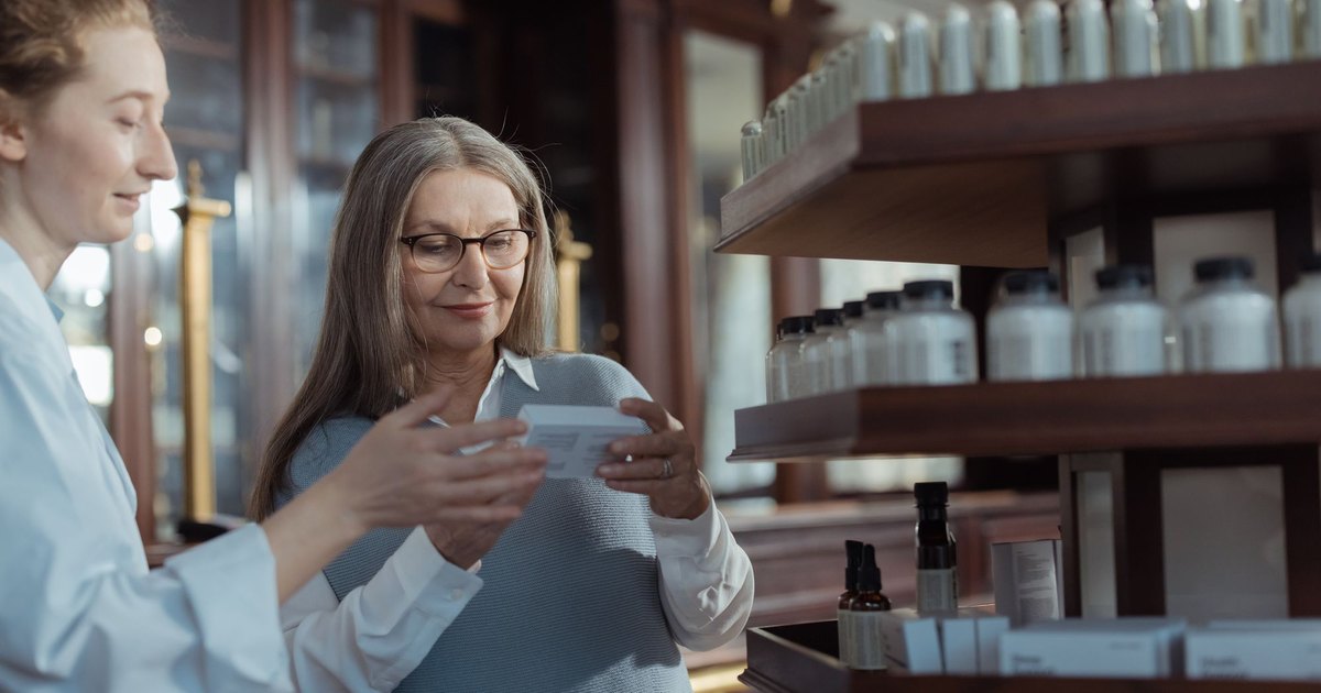 Older woman reading a small box while a pharmacist in a white coat helps her at the counter
