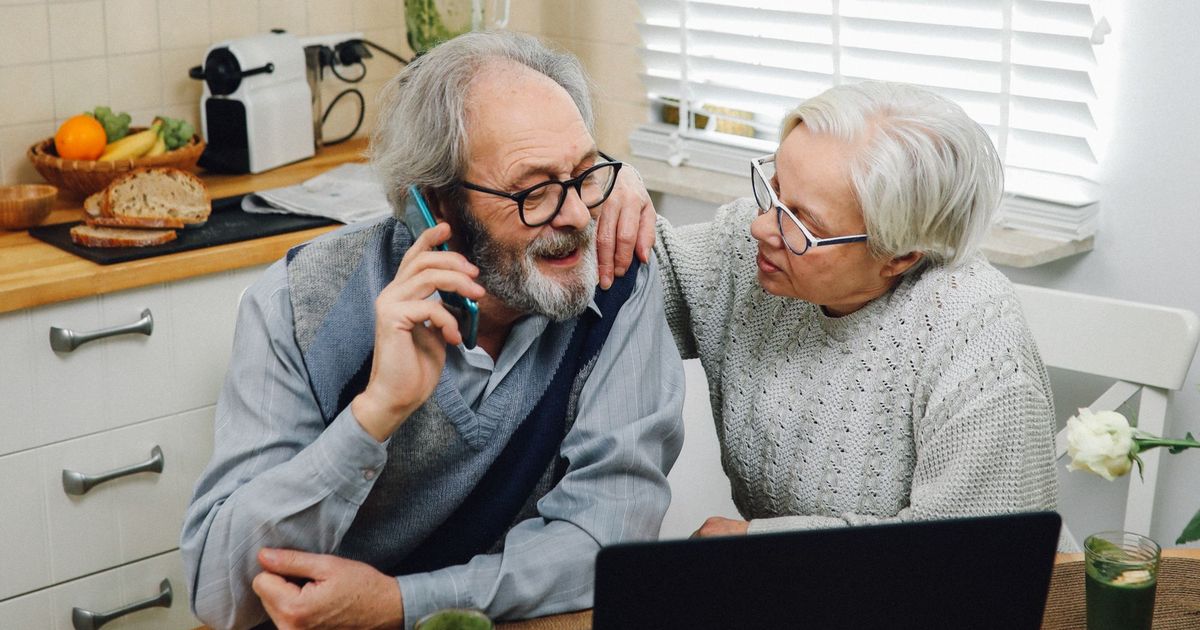 Older couple at a kitchen table with a laptop, one of them on the phone