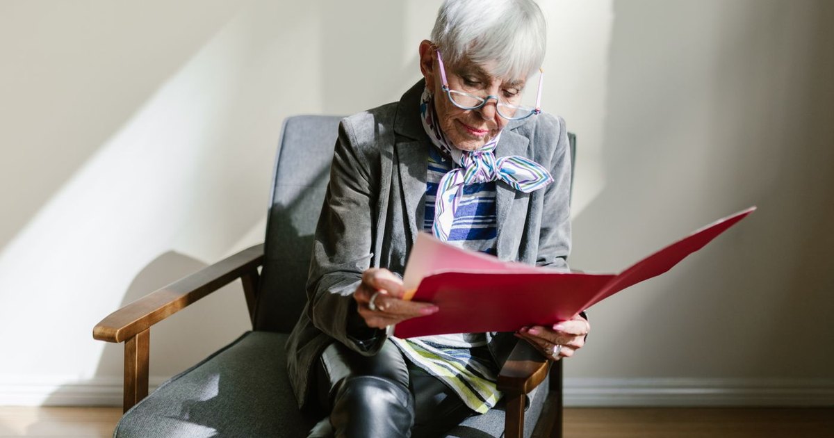 Older woman with reading glasses seated in an armchair, reading a document from a red folder